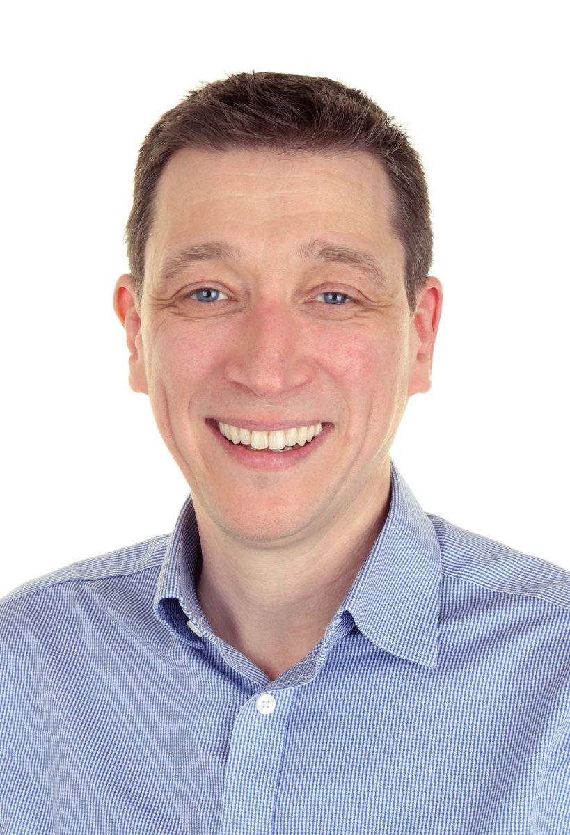 Smiling Mark from DIS Consulting IT wearing a light blue checkered shirt, standing against a plain white background.