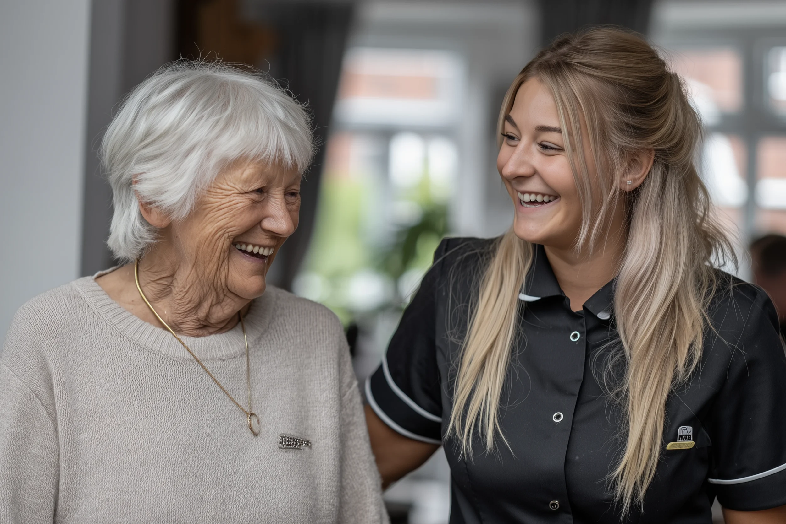 Elderly woman smiling warmly alongside a young care worker in a homely care setting.