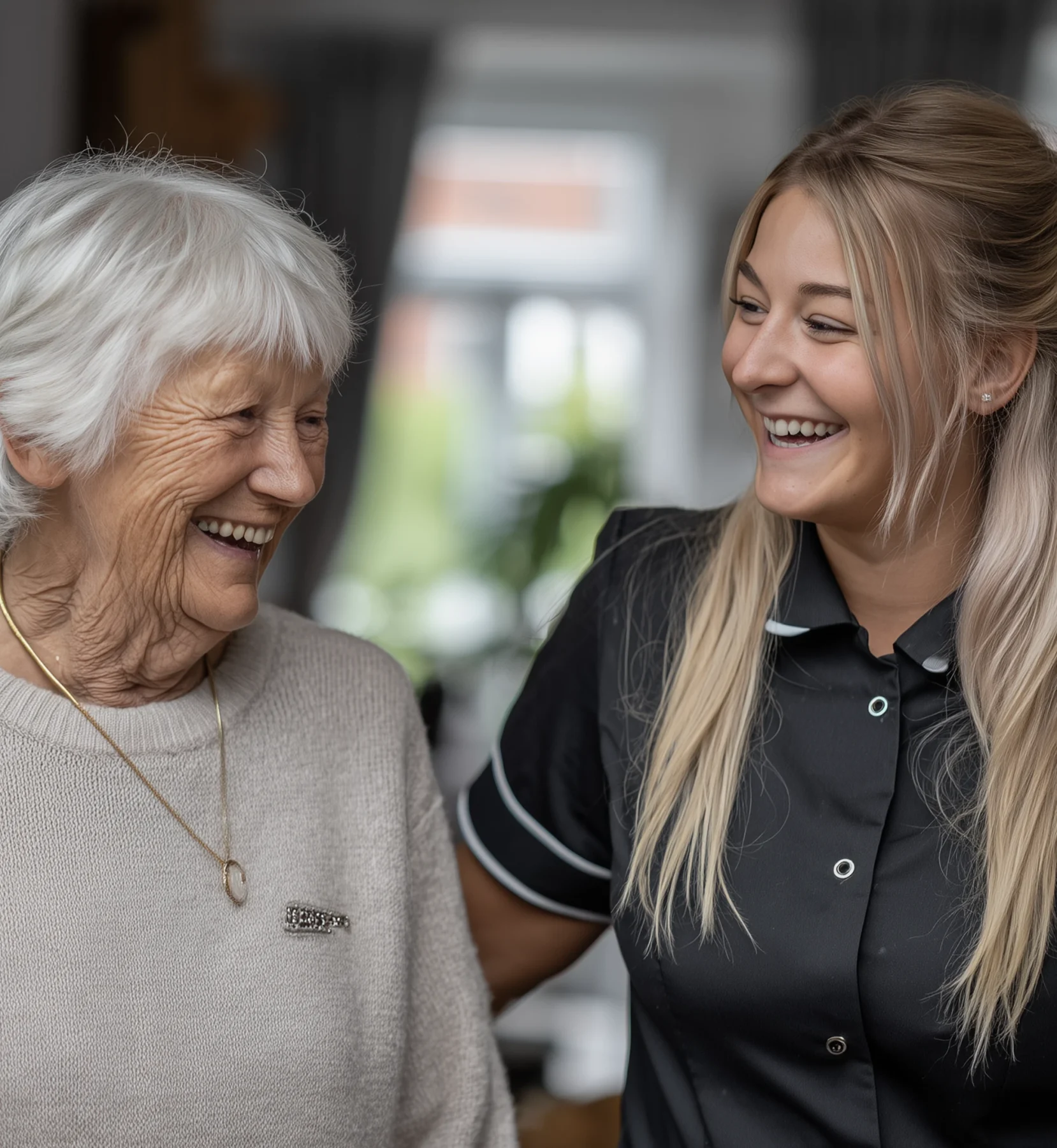 Elderly woman smiling warmly alongside a young care worker in a homely care setting.