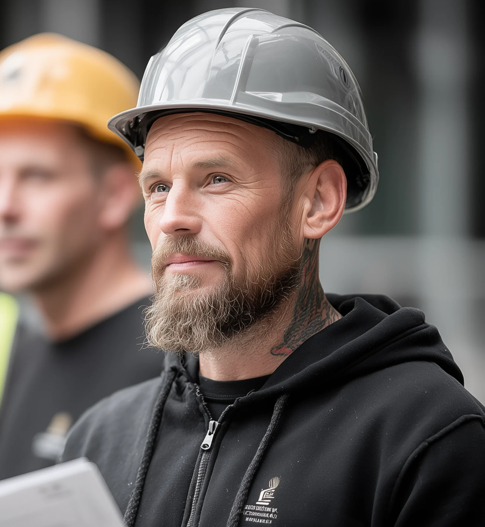 Construction worker wearing a safety helmet, standing on-site and looking confident while holding documents.