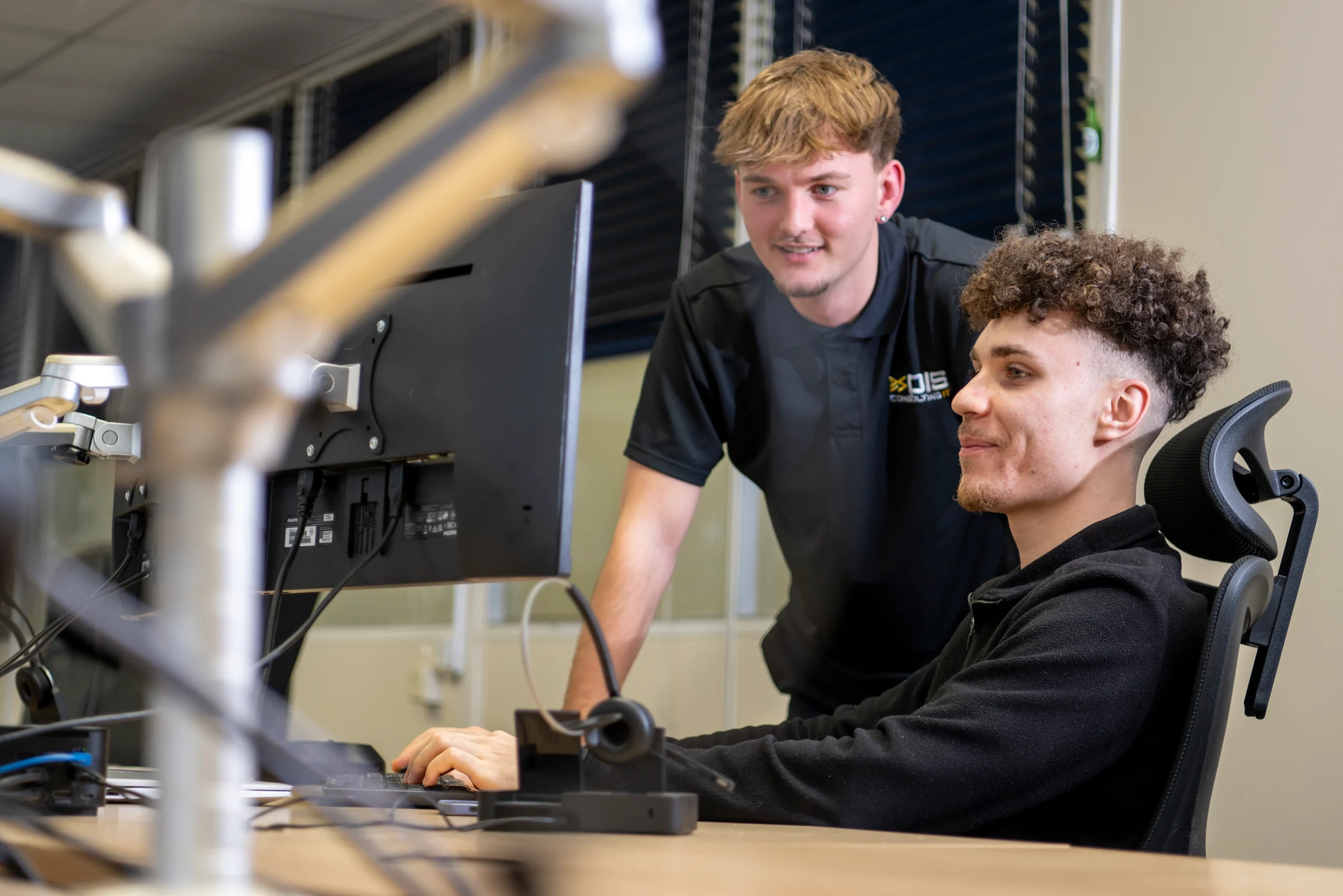 Scott assisting Ethan at his desk in the DISConsulting IT support office, both focused on resolving a technical issue together.